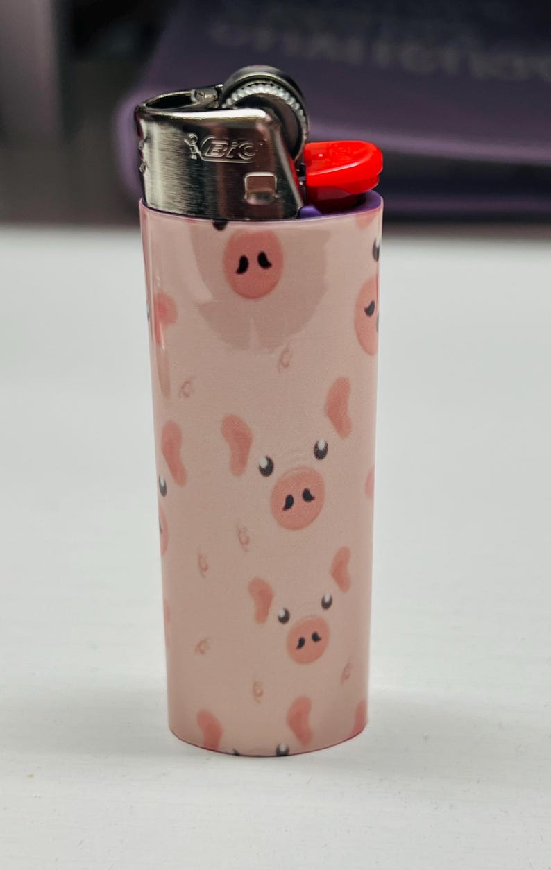 K&ouml;nnte beinhalten: Ein rosa BIC-Feuerzeug mit einem sich wiederholenden Muster aus Cartoon-Schweinegesichtern. Das Feuerzeug hat einen silbernen Metallaufsatz und einen roten Z&uuml;ndknopf. Die Schweinegesichter haben schwarze Nasenl&ouml;cher und kleine, gebogene Ohren.