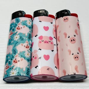 K&ouml;nnte beinhalten: Drei neuartige Feuerzeuge mit Schweinedesigns. Ein Feuerzeug hat einen t&uuml;rkisfarbenen Hintergrund mit rosa Schweinen und wei&szlig;en Blumen. Das zweite Feuerzeug hat einen wei&szlig;en Hintergrund mit rosa Schweinen und Herzen. Das dritte Feuerzeug hat einen rosa Hintergrund mit Schweinegesichtern.
