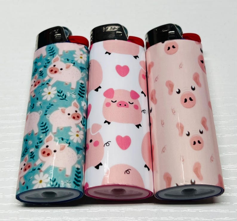 K&ouml;nnte beinhalten: Drei neuartige Feuerzeuge mit Schweinedesigns. Ein Feuerzeug hat einen t&uuml;rkisfarbenen Hintergrund mit rosa Schweinen und wei&szlig;en Blumen. Das zweite Feuerzeug hat einen wei&szlig;en Hintergrund mit rosa Schweinen und Herzen. Das dritte Feuerzeug hat einen rosa Hintergrund mit Schweinegesichtern.
