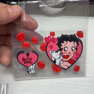 Op de afbeelding: Een doorzichtige plastic zak met een verzameling stickers met het thema Betty Boop. De set bevat een grote hartvormige sticker met Betty Boop en haar hond, kleinere hartvormige stickers met de tekst Betty Boop en diverse rode lippenvormige stickers.