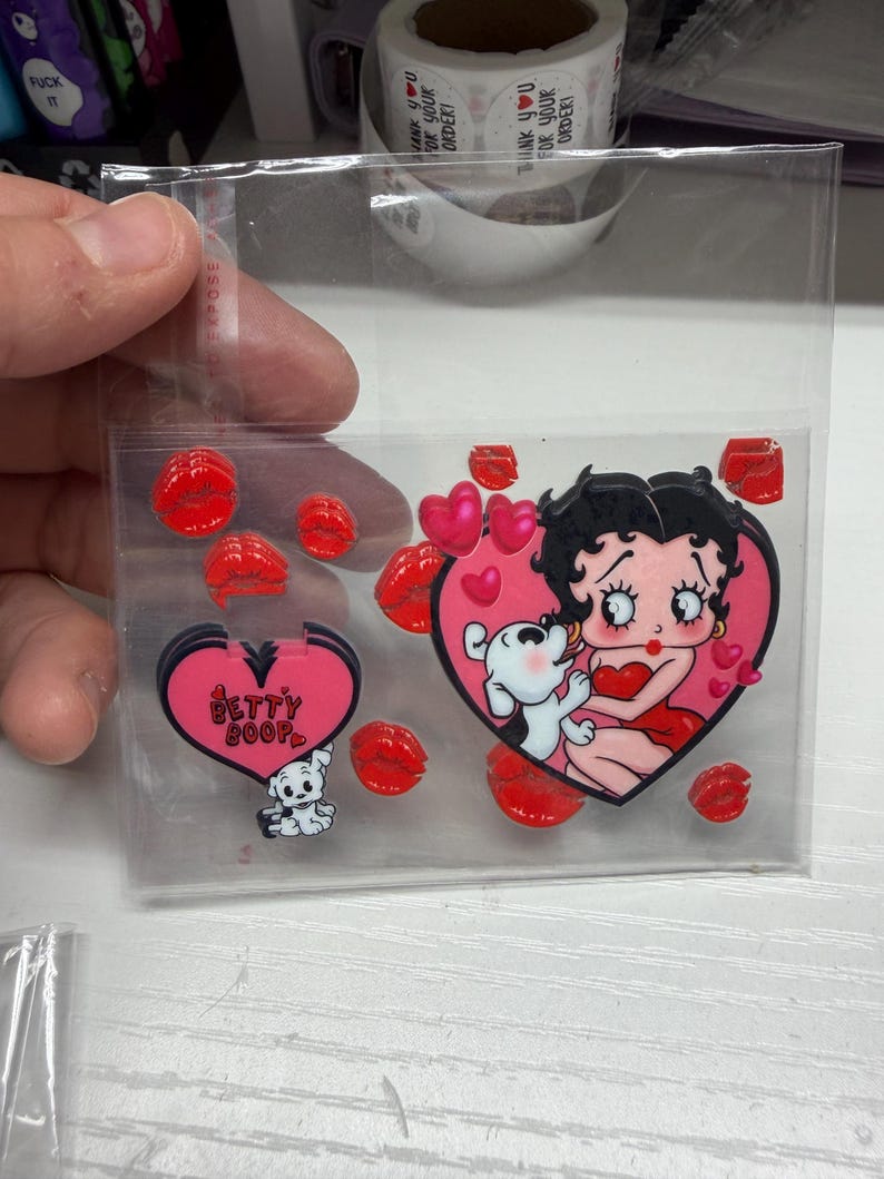 Op de afbeelding: Een doorzichtige plastic zak met een verzameling stickers met het thema Betty Boop. De set bevat een grote hartvormige sticker met Betty Boop en haar hond, kleinere hartvormige stickers met de tekst Betty Boop en diverse rode lippenvormige stickers.