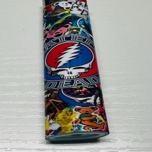 Puede incluir: Un encendedor colorido con las palabras "STEAL YOUR FACE" y "DEAD" con un dise&ntilde;o de calavera y rayo. El encendedor tambi&eacute;n tiene un gr&aacute;fico de oso de dibujos animados y otras ilustraciones coloridas.