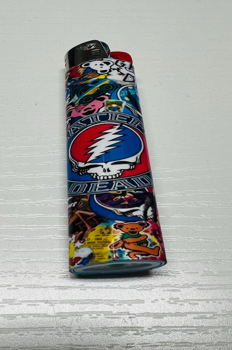 Puede incluir: Un encendedor colorido con las palabras "STEAL YOUR FACE" y "DEAD" con un dise&ntilde;o de calavera y rayo. El encendedor tambi&eacute;n tiene un gr&aacute;fico de oso de dibujos animados y otras ilustraciones coloridas.