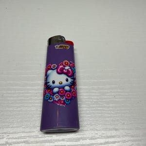 以下が含まれることがあります： ハローキティのデザインが施された紫色のBICライター。キャラクターの周りには花のハートのデザインがあります。ライターの上部は黒で、赤い点火ボタンが付いています。