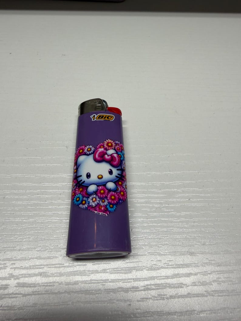以下が含まれることがあります： ハローキティのデザインが施された紫色のBICライター。キャラクターの周りには花のハートのデザインがあります。ライターの上部は黒で、赤い点火ボタンが付いています。