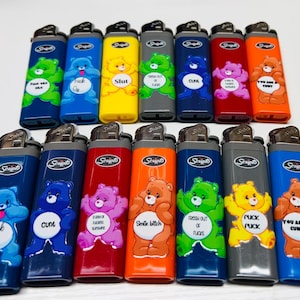 May include: A collection of novelty lighters in various colors, each featuring a cartoon bear design with bold text. The lighters are blue, green, yellow, gray, red, and orange. The text includes phrases like "Fuck Out", "Slut", and "You Are a Cunt".