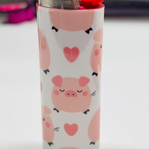 K&ouml;nnte beinhalten: Ein wei&szlig;es Feuerzeug mit einem sich wiederholenden Muster aus rosa Schweinen und Herzen. Das Feuerzeug hat einen silbernen Metallaufsatz und einen roten Knopf. Das Design zeigt Cartoon-Schweine mit geschlossenen Augen und kleinen Lockenschw&auml;nzen.