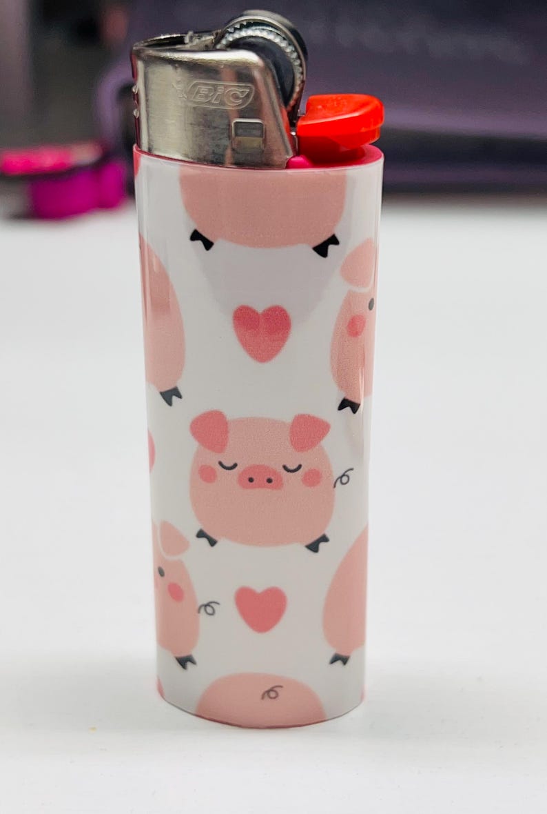K&ouml;nnte beinhalten: Ein wei&szlig;es Feuerzeug mit einem sich wiederholenden Muster aus rosa Schweinen und Herzen. Das Feuerzeug hat einen silbernen Metallaufsatz und einen roten Knopf. Das Design zeigt Cartoon-Schweine mit geschlossenen Augen und kleinen Lockenschw&auml;nzen.