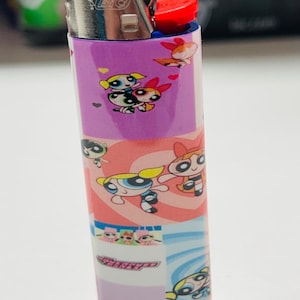 May include: A purple BIC lighter decorated with Powerpuff Girls cartoon characters. The lighter has a red ignition button and a silver metal top. The design includes images of Blossom, Bubbles, and Buttercup.