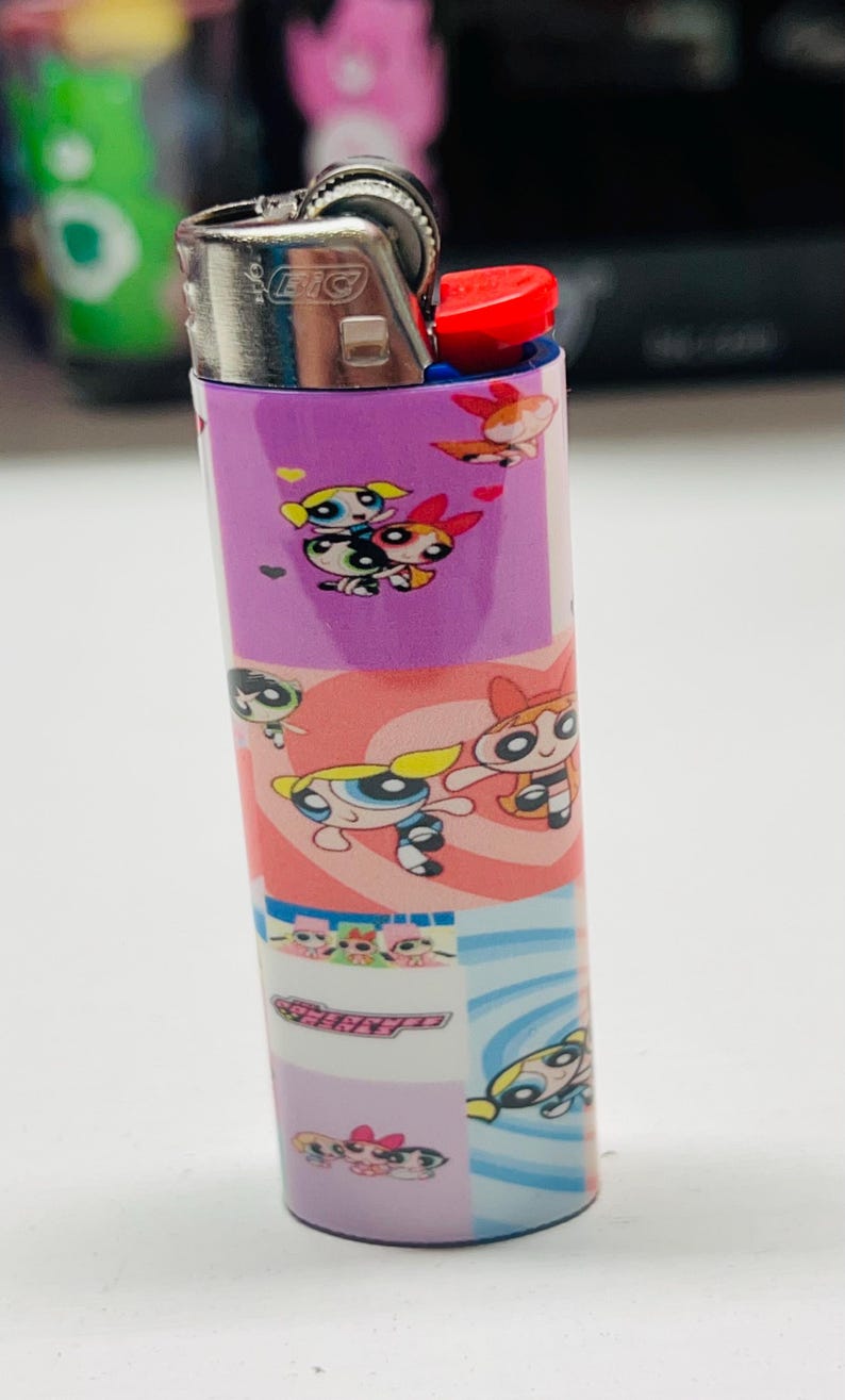 May include: A purple BIC lighter decorated with Powerpuff Girls cartoon characters. The lighter has a red ignition button and a silver metal top. The design includes images of Blossom, Bubbles, and Buttercup.