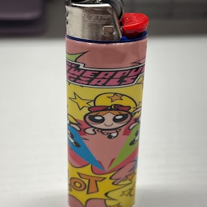 May include: A BIC lighter with a vibrant design showcasing the text "POWERPUFF GIRLS" and cartoon characters. The lighter has a silver metal top with a red ignition button and a pink, yellow, and blue patterned body.