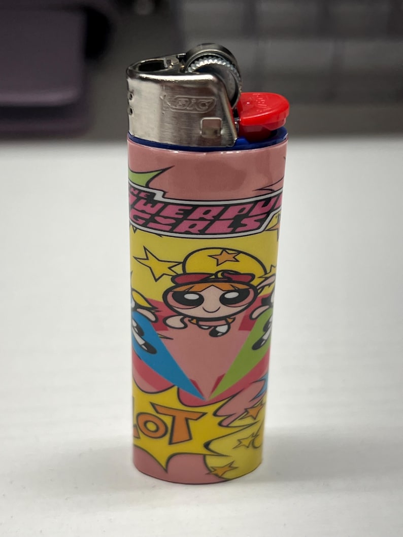May include: A BIC lighter with a vibrant design showcasing the text "POWERPUFF GIRLS" and cartoon characters. The lighter has a silver metal top with a red ignition button and a pink, yellow, and blue patterned body.