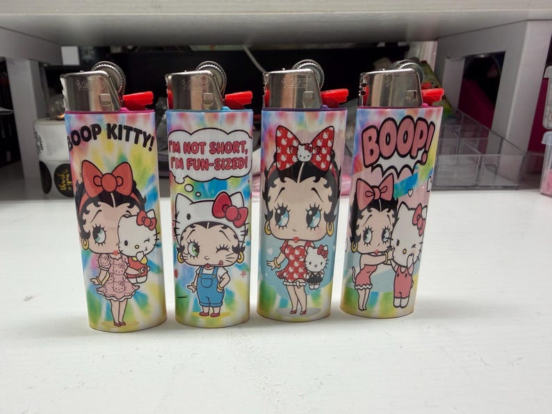 May include: Four cylindrical lighters featuring a colorful tie-dye background and cartoon graphics of Betty Boop and Hello Kitty. The lighters display text including BOOP KITTY, I'M NOT SHORT, I'M FUN-SIZED, and BOOP, with metallic tops.