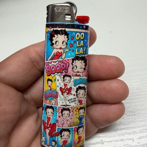 May include: A rectangular lighter featuring a multi-panel comic strip design of Betty Boop in various poses and outfits. The graphic includes text bubbles with phrases like OO LA LA!, BOOP!, SWOON, YOWZA!, CHEEKY, CLASSIC, and SPARKLE. The lighter has a silver metal top.