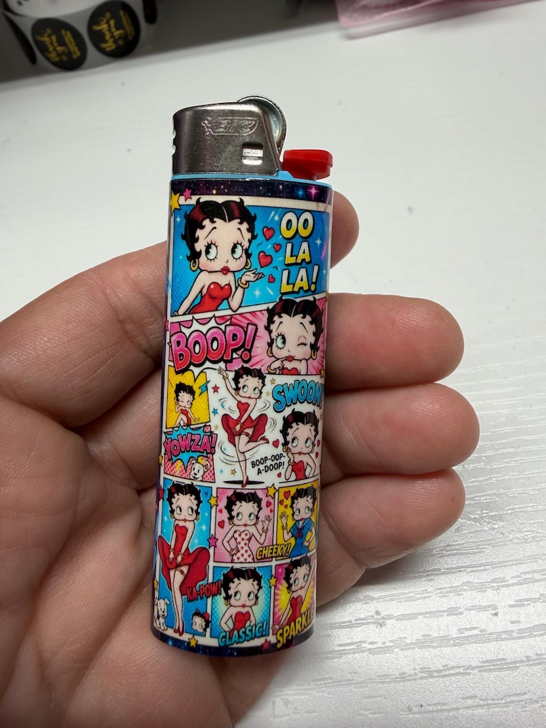 May include: A rectangular lighter featuring a multi-panel comic strip design of Betty Boop in various poses and outfits. The graphic includes text bubbles with phrases like OO LA LA!, BOOP!, SWOON, YOWZA!, CHEEKY, CLASSIC, and SPARKLE. The lighter has a silver metal top.
