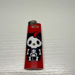 以下が含まれることがあります： ハローキティのスケルトンデザインが施された赤いBICライター。ライターには、白黒のスケルトン柄と赤いリボンが描かれています。BICのロゴがライターの上部に表示されています。