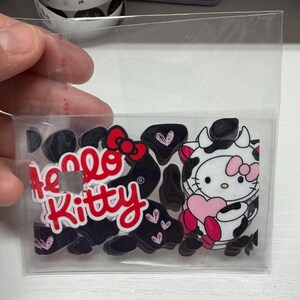 Op de afbeelding: Een doorzichtig plastic zakje met meerdere acryl bedels met het Hello Kitty logo in rode tekst en een personage in een zwart-wit koeienprint outfit dat een roze hart vasthoudt, omringd door kleine zwarte en roze hartvormige stukjes.