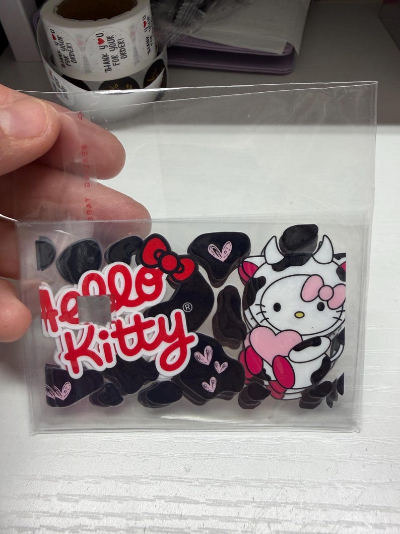 Op de afbeelding: Een doorzichtig plastic zakje met meerdere acryl bedels met het Hello Kitty logo in rode tekst en een personage in een zwart-wit koeienprint outfit dat een roze hart vasthoudt, omringd door kleine zwarte en roze hartvormige stukjes.