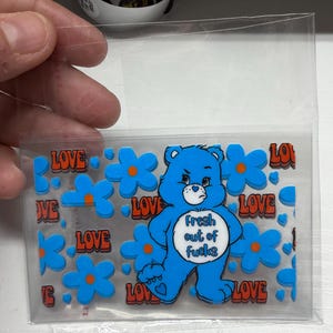 Op de afbeelding: Een rechthoekig, doorzichtig stickervel met een blauwe beer met een wit buikembleem waarop de tekst Fresh out of fucks staat. Het ontwerp bevat herhalende blauwe bloemen met oranje harten en het woord LOVE in oranje letters.