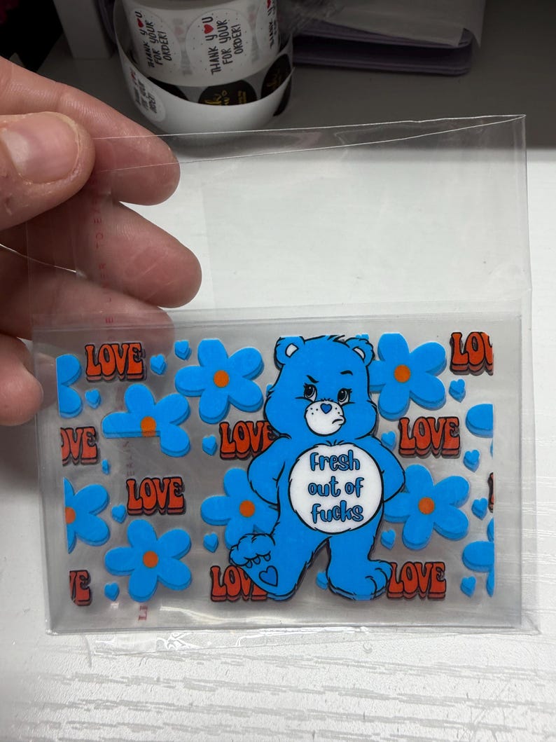 Op de afbeelding: Een rechthoekig, doorzichtig stickervel met een blauwe beer met een wit buikembleem waarop de tekst Fresh out of fucks staat. Het ontwerp bevat herhalende blauwe bloemen met oranje harten en het woord LOVE in oranje letters.