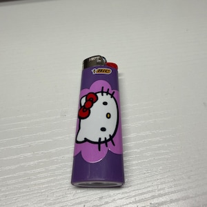 以下が含まれることがあります： ハローキティのデザインが施された紫色のBICライター。ライターには、赤いリボン、黒い目、ひげが付いた白いハローキティの顔があります。ライターは白い表面にあります。