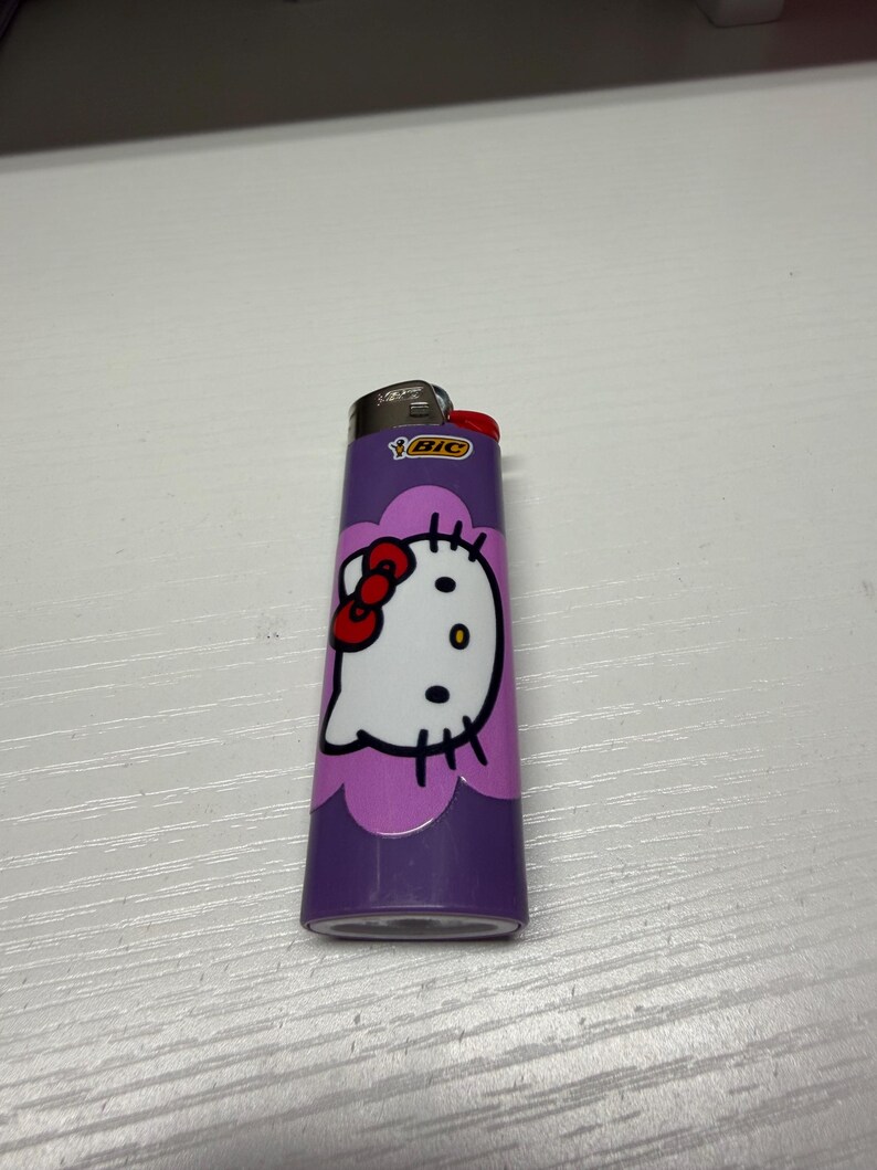 以下が含まれることがあります： ハローキティのデザインが施された紫色のBICライター。ライターには、赤いリボン、黒い目、ひげが付いた白いハローキティの顔があります。ライターは白い表面にあります。