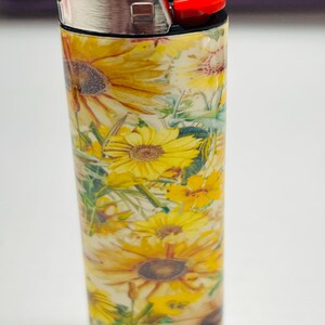 Pode incluir: Um isqueiro com um padr&atilde;o floral com girass&oacute;is amarelos e folhas verdes. A parte superior &eacute; de metal prateado, com um bot&atilde;o de igni&ccedil;&atilde;o vermelho. O design &eacute; brilhante e alegre.