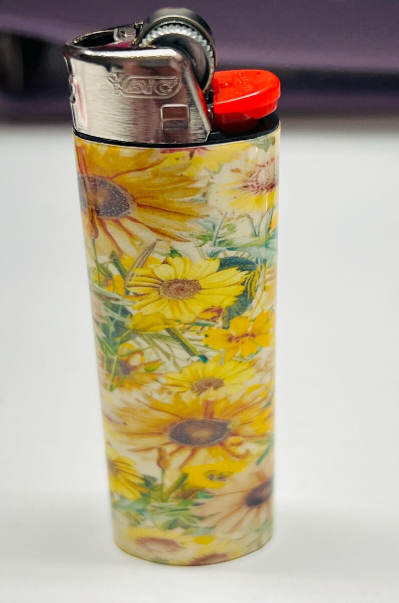 Pode incluir: Um isqueiro com um padr&atilde;o floral com girass&oacute;is amarelos e folhas verdes. A parte superior &eacute; de metal prateado, com um bot&atilde;o de igni&ccedil;&atilde;o vermelho. O design &eacute; brilhante e alegre.