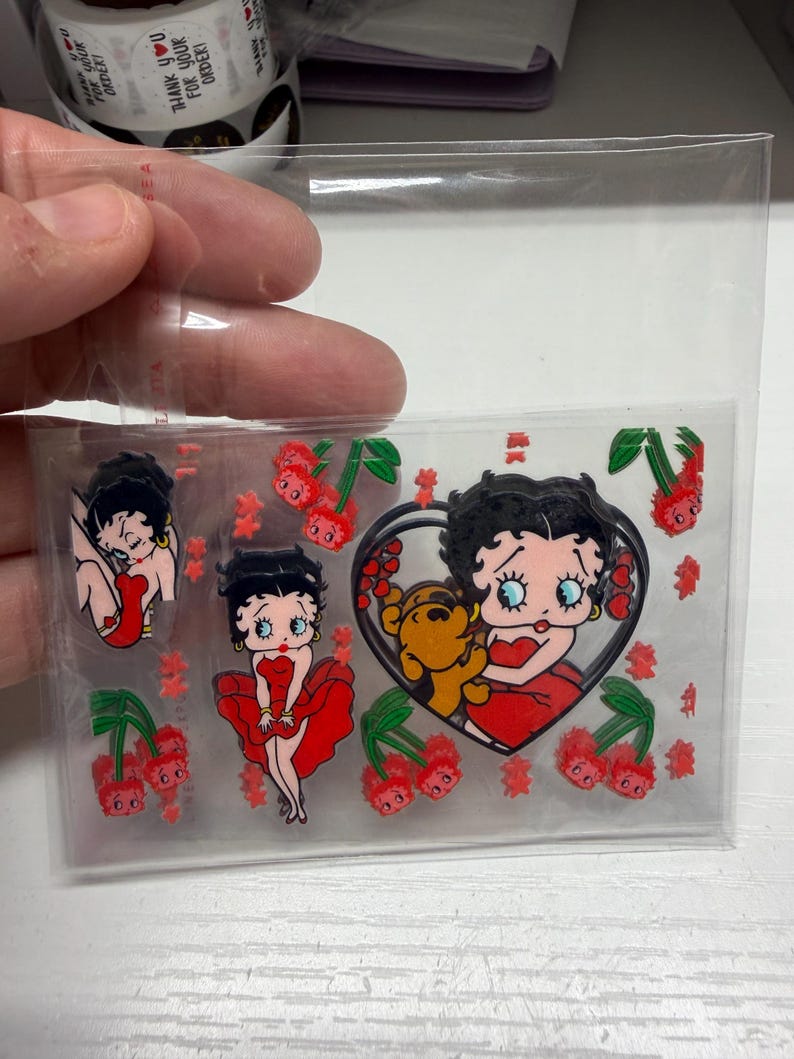 Op de afbeelding: Een doorzichtig plastic vel met meerdere stickers van een tekenfilmfiguur met zwart haar in een rode jurk, een bruine hond en rode kersen. De stickers zijn gerangschikt op een transparante ondergrond en tonen diverse houdingen.