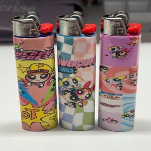 May include: Three BIC lighters with Powerpuff Girls designs. The lighters feature the characters Blossom, Bubbles, and Buttercup. Each lighter has a different colour scheme, including pink, blue, and purple, with the text "POWERPUFF GIRLS".