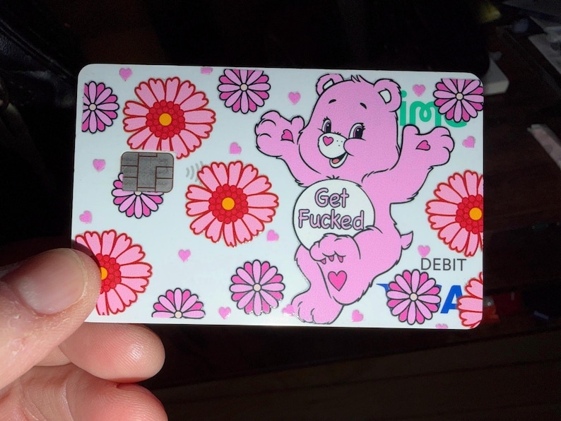 Op de afbeelding: Een witte betaalpas met een herhalend patroon van roze en rode bloemen, kleine roze hartjes en een roze cartoonbeer met een cirkelvormig embleem op de borst met de tekst Get Fucked in zwarte letters, voorzien van een zilverkleurige chip.