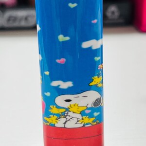 May include: A blue BIC lighter featuring a Snoopy and Woodstock design. The lighter has a red ignition button and a silver metal top. The Snoopy graphic is set against a blue sky background with colorful hearts and clouds.