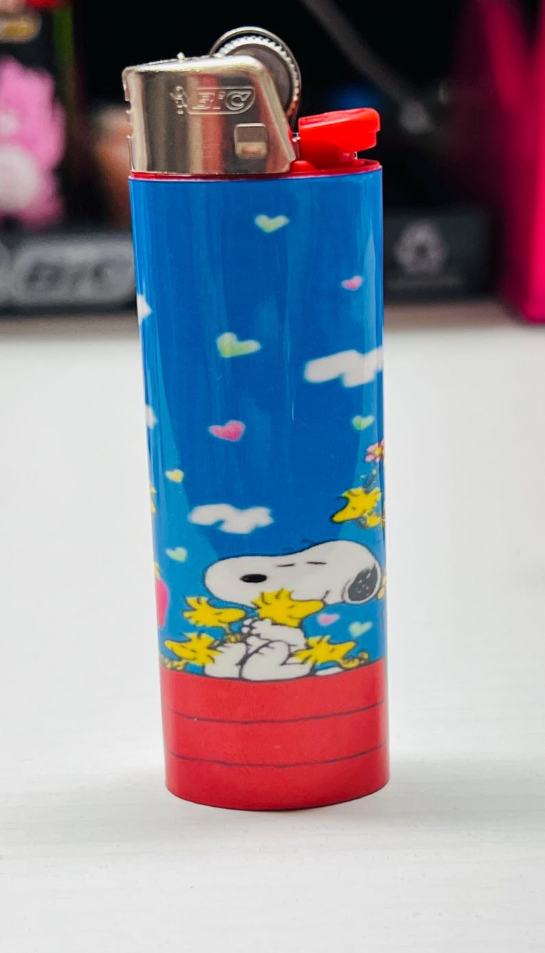 May include: A blue BIC lighter featuring a Snoopy and Woodstock design. The lighter has a red ignition button and a silver metal top. The Snoopy graphic is set against a blue sky background with colorful hearts and clouds.