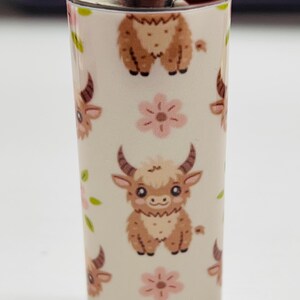 Pode incluir: Um isqueiro BIC branco com um padr&atilde;o repetido de vacas das Terras Altas de desenho animado e flores cor-de-rosa. O isqueiro tem um bot&atilde;o de igni&ccedil;&atilde;o vermelho e a marca "BIC" &eacute; vis&iacute;vel.