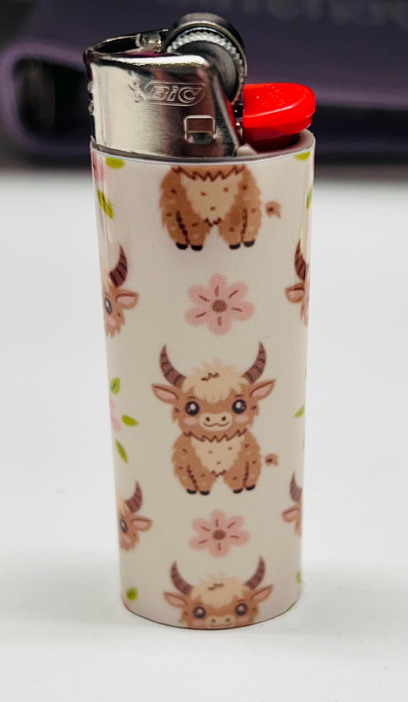 Pode incluir: Um isqueiro BIC branco com um padr&atilde;o repetido de vacas das Terras Altas de desenho animado e flores cor-de-rosa. O isqueiro tem um bot&atilde;o de igni&ccedil;&atilde;o vermelho e a marca "BIC" &eacute; vis&iacute;vel.