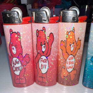 Puede incluir: Tres encendedores cubiertos de purpurina con ilustraciones al estilo Care Bear. Los encendedores presentan osos rojos, naranjas y rosas con frases como "Who Cares" y "Wish A Bitch Would". Cada encendedor tiene una tapa de metal plateado y detalles en rojo.