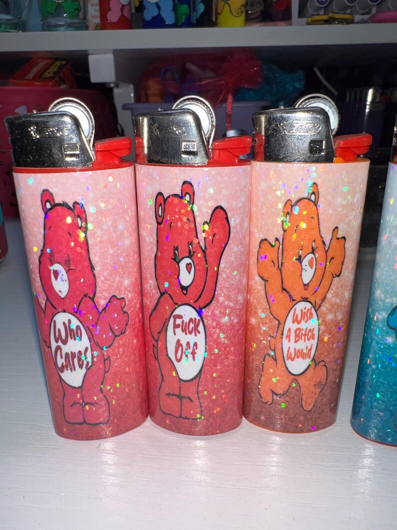 Puede incluir: Tres encendedores cubiertos de purpurina con ilustraciones al estilo Care Bear. Los encendedores presentan osos rojos, naranjas y rosas con frases como "Who Cares" y "Wish A Bitch Would". Cada encendedor tiene una tapa de metal plateado y detalles en rojo.