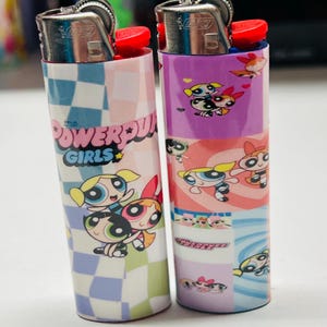 May include: Two lighters with cartoon character designs. One has a blue and white chequered pattern with the text "The Powerpuff Girls". The other has a purple and pink design featuring the characters from the show.