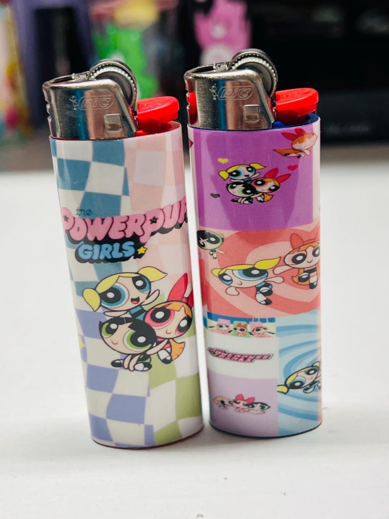 May include: Two lighters with cartoon character designs. One has a blue and white chequered pattern with the text "The Powerpuff Girls". The other has a purple and pink design featuring the characters from the show.
