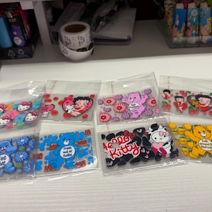 Op de afbeelding: Een verzameling van acht doorzichtige plastic zakjes met diverse stickers met karakterthema's. De ontwerpen bevatten Hello Kitty, Betty Boop en kleurrijke beren omringd door bloemen, harten en kersen, waarvan sommige met tekst zoals Hello Kitty.