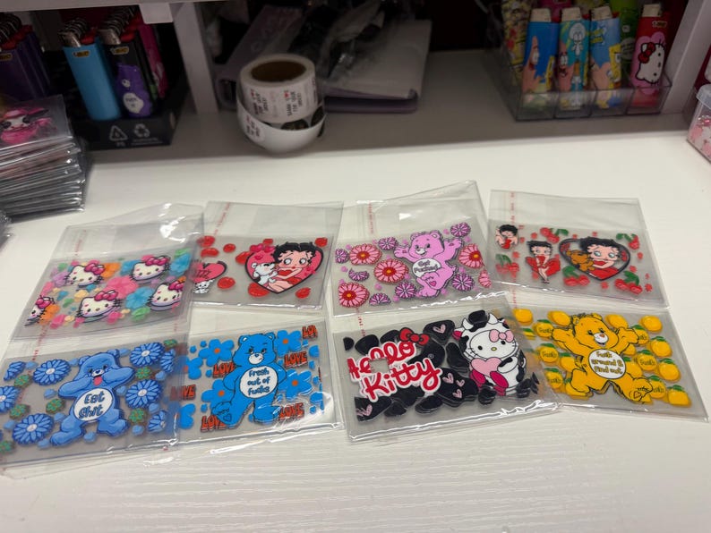 Op de afbeelding: Een verzameling van acht doorzichtige plastic zakjes met diverse stickers met karakterthema's. De ontwerpen bevatten Hello Kitty, Betty Boop en kleurrijke beren omringd door bloemen, harten en kersen, waarvan sommige met tekst zoals Hello Kitty.