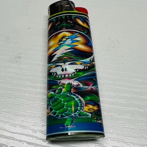 Puede incluir: Un encendedor colorido con un dise&ntilde;o vibrante que presenta una calavera, un rayo y una tortuga verde. El encendedor tiene una parte superior negra con un bot&oacute;n de encendido rojo y una base azul.