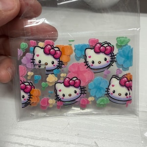 Op de afbeelding: Een doorzichtig plastic zakje met meerdere platte, decoratieve Hello Kitty-figuren met roze strikjes, afgewisseld met diverse kleine, kleurrijke bloemvormige confetti in de kleuren blauw, groen, oranje en roze.