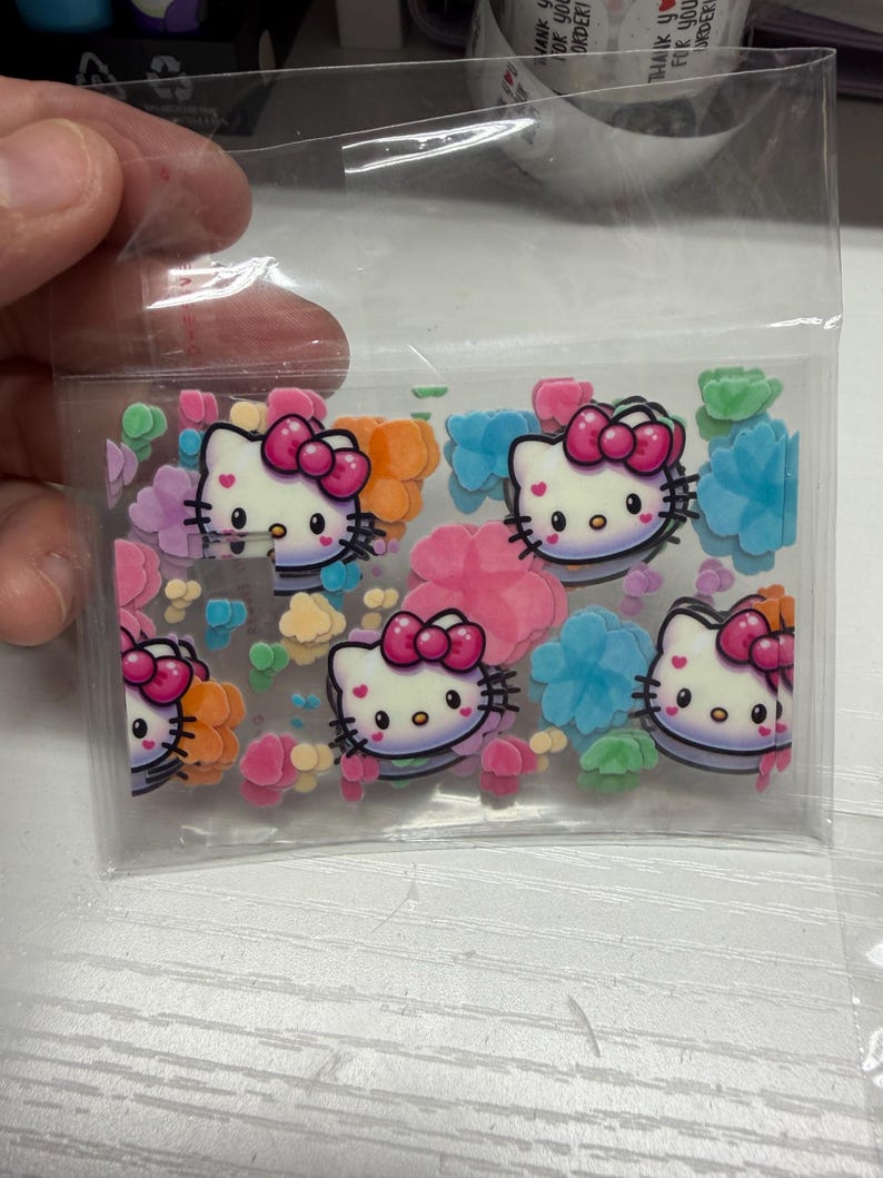 Op de afbeelding: Een doorzichtig plastic zakje met meerdere platte, decoratieve Hello Kitty-figuren met roze strikjes, afgewisseld met diverse kleine, kleurrijke bloemvormige confetti in de kleuren blauw, groen, oranje en roze.