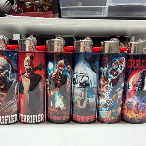 May include: Six cylindrical lighters arranged in a row, each featuring a unique graphic print of the character Art the Clown from the Terrifier film series. Each lighter displays the word Terrifier in white or red text at the base of the design.