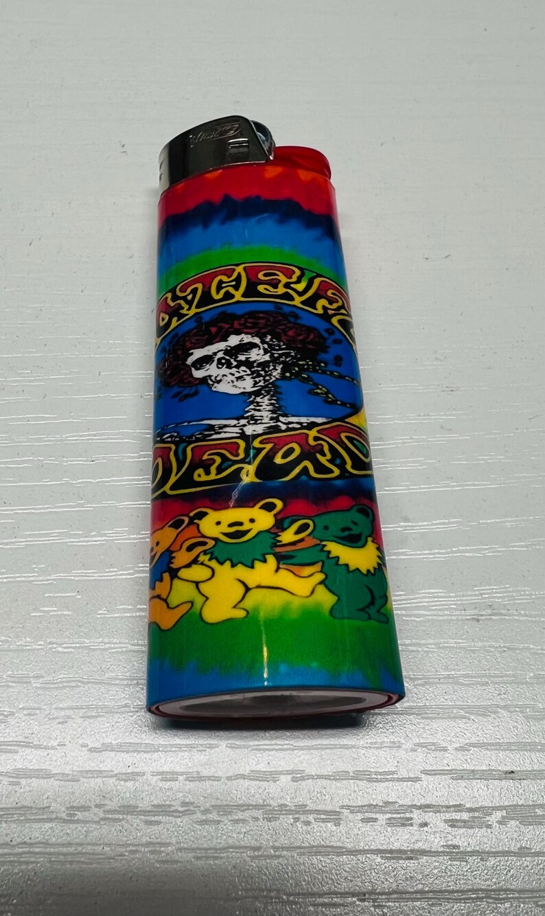 Puede incluir: Un encendedor colorido con un dise&ntilde;o tie-dye en la parte superior y una vibrante ilustraci&oacute;n de una calavera y osos. El encendedor tiene una parte superior de metal y un cuerpo verde, azul y amarillo.