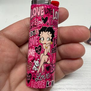 May include: A pink Bic lighter featuring a repeating pattern of the word LOVE in various fonts, hearts, and a graphic illustration of the character Betty Boop with her dog. The design includes the text Boop-o-oop and Betty Boop in black and white.