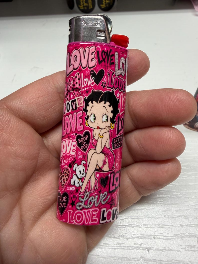 May include: A pink Bic lighter featuring a repeating pattern of the word LOVE in various fonts, hearts, and a graphic illustration of the character Betty Boop with her dog. The design includes the text Boop-o-oop and Betty Boop in black and white.