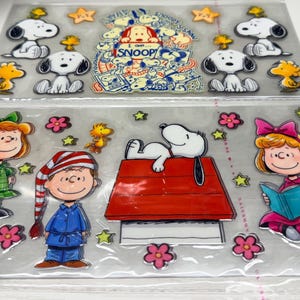 May include: Two clear sheets of Peanuts character stickers. The top sheet features Snoopy and Woodstock in various poses with stars. The bottom sheet displays Snoopy on a red doghouse, Charlie Brown in blue pajamas, and two girls with books.