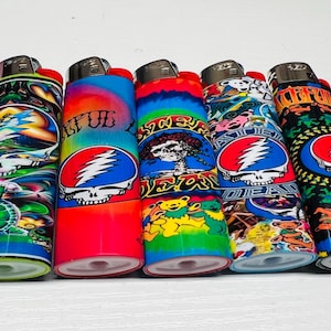 Puede incluir: Cinco encendedores coloridos con varios dise&ntilde;os, incluyendo una tortuga, una calavera con rayos y osos bailarines. Los encendedores tienen botones de encendido rojos y est&aacute;n sobre una superficie blanca.